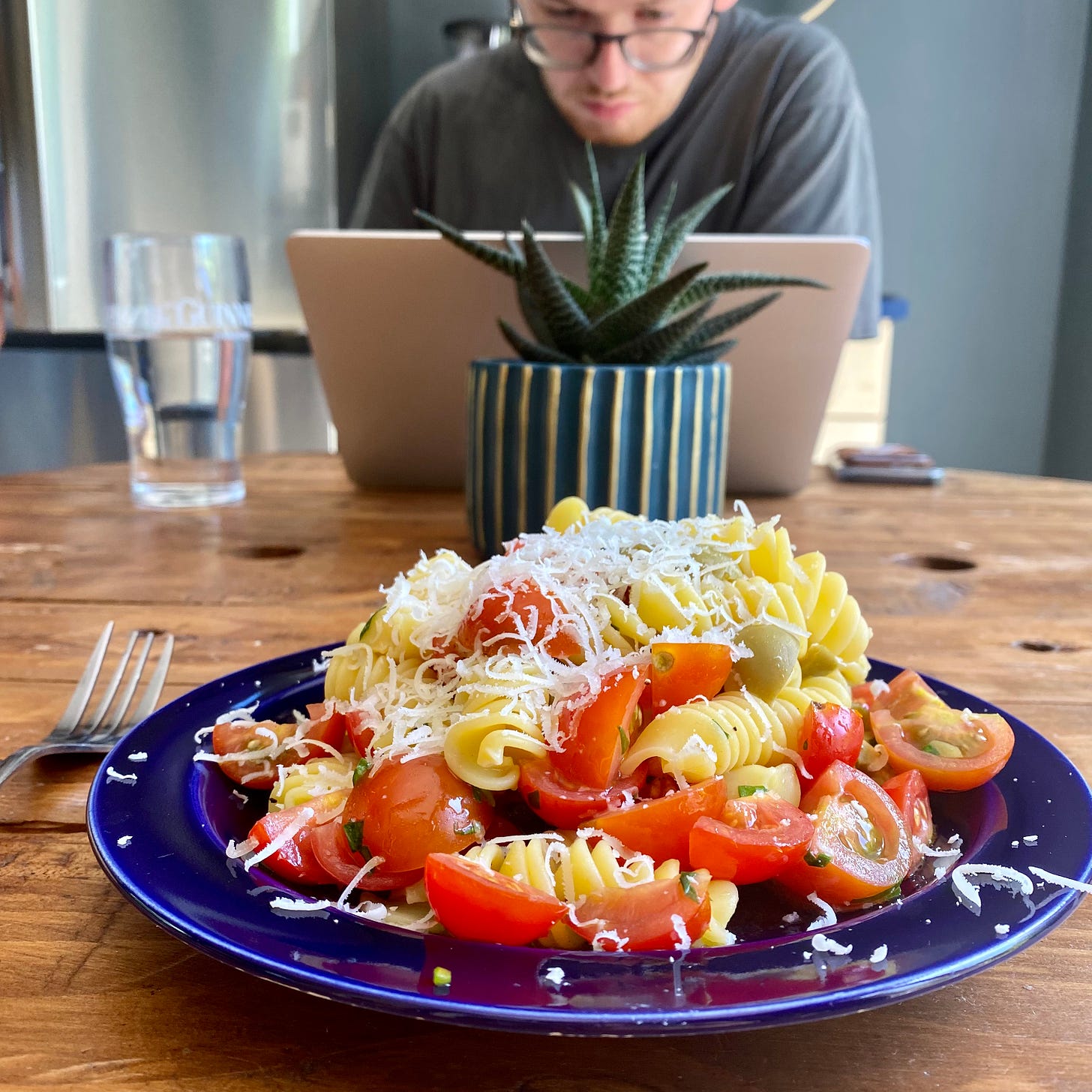 Blue plate serving fusilli pasta with fresh tomatoes and green olives, scattered with grated cheese. Person working at a laptop in the background.