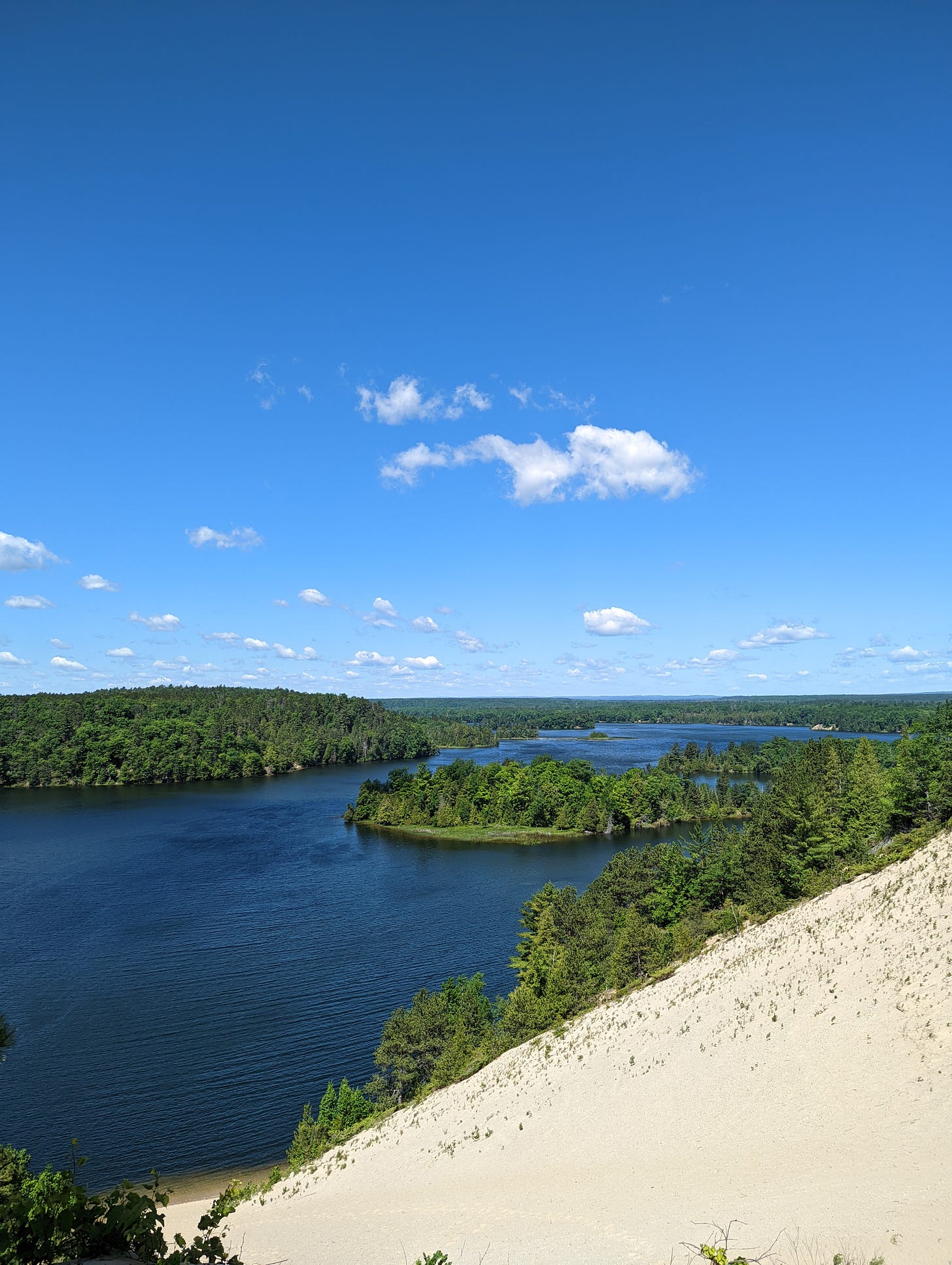 A few clouds hang on a bright blue sky above a finger lake and bright green forest trees. A sand dune is in the foreground.