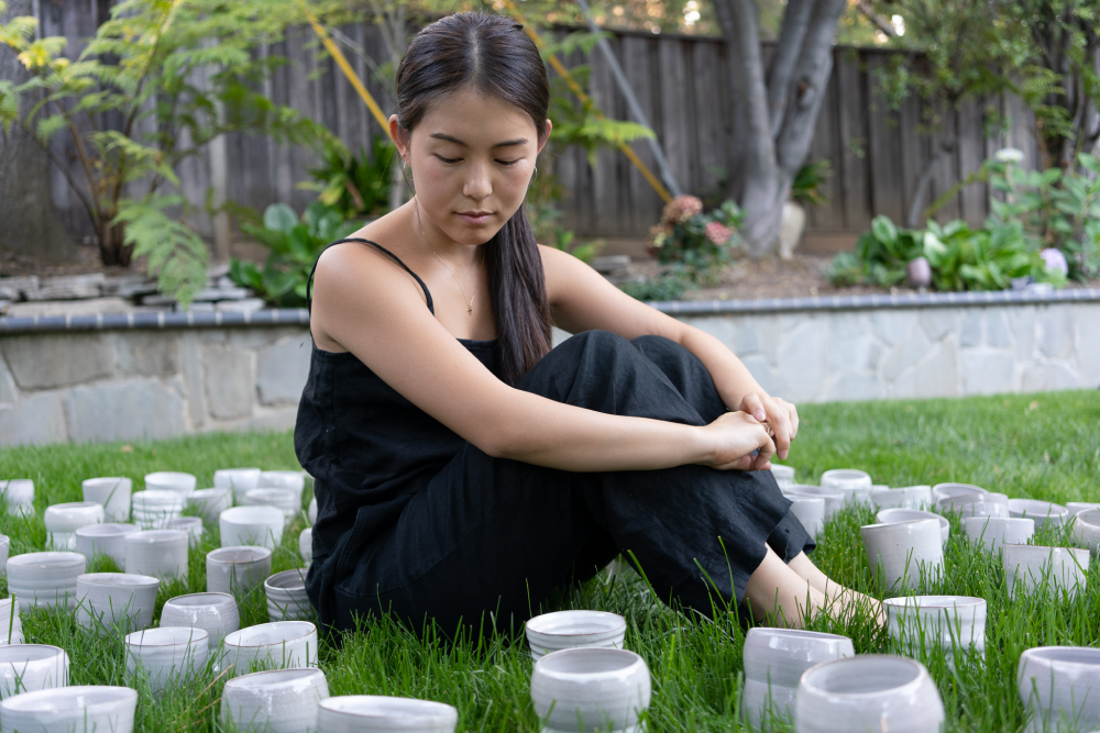 Janice sitting on green grass with her arms around her legs, looking down at ceramic tumblers she made for her wedding guests.