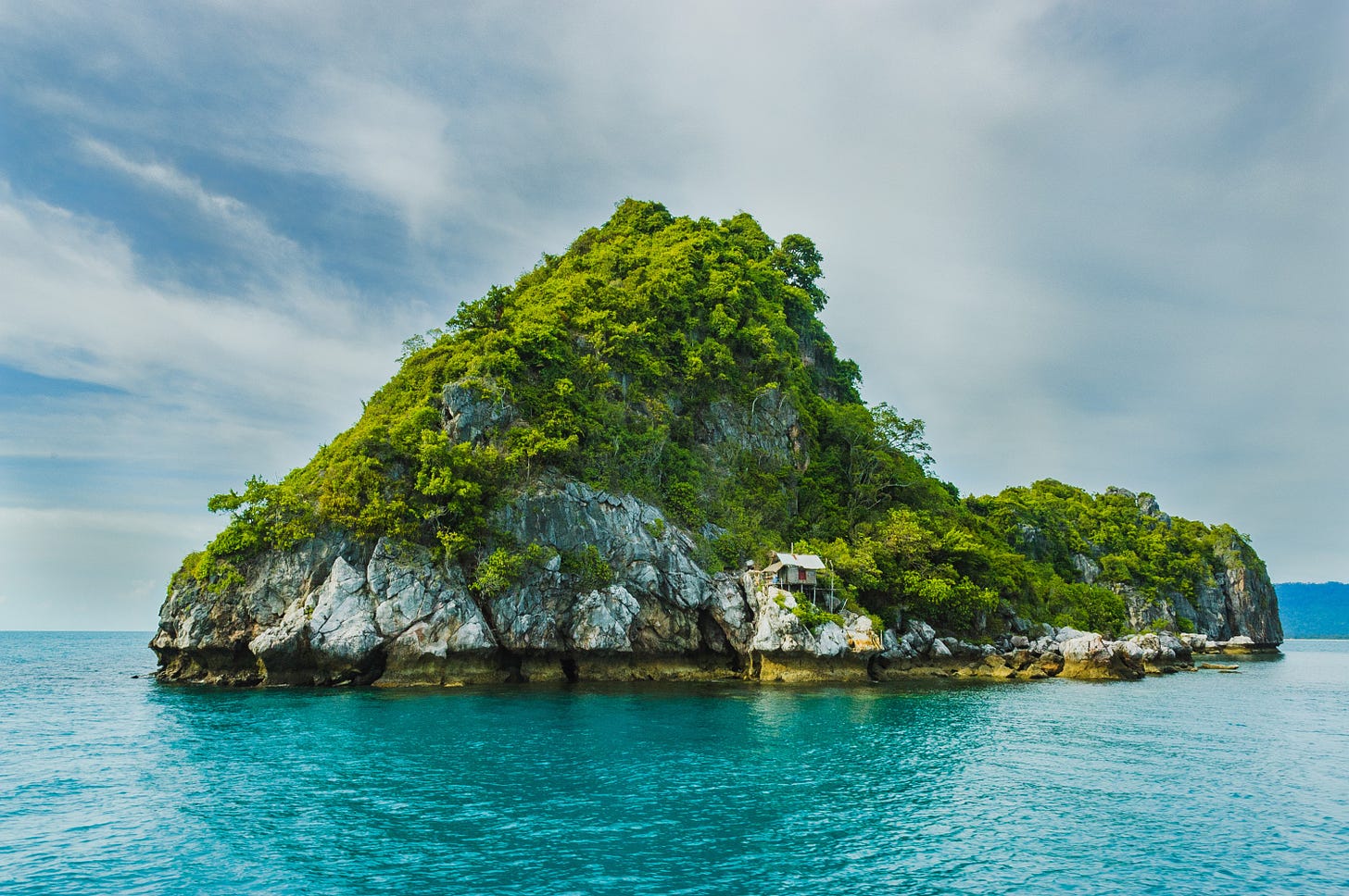 A rocky outcrop sits as a small island in clear blue water. A small hut sits along the shore of the mountain, nestled beside the rock face.