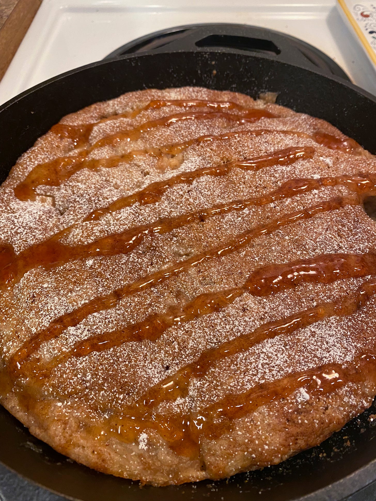 In a cast iron pan, a golden-brown apple cake. On top is a drizzle of syrup and a dusting of powdered sugar.