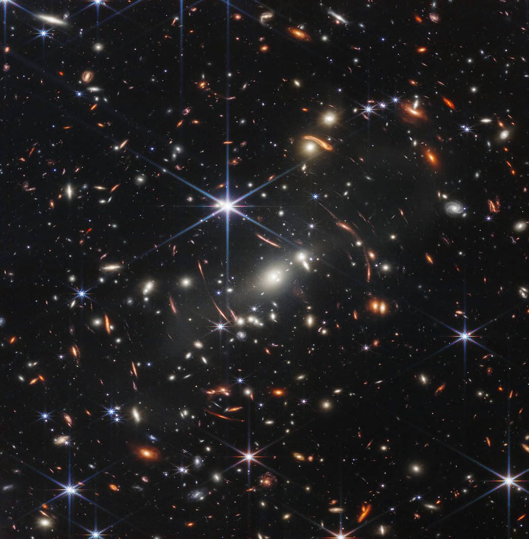 distant galaxies appear as bright glowing spots in this Webb telescope image, with some smeared by gravitational lensing; foreground stars appear bright with six-pointed diffraction spikes, owing to the shape of Webb's mirrors