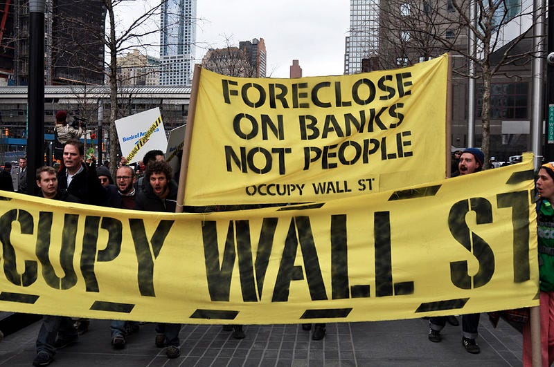 Photograph of a protest rally in the US, foregrounding two yellow banners. The banner on top is small and has text which says “Foreclose on banks, not people” & “Occupy wall street”. The second banner is longer and it reads “Occupy Wall Street”.
