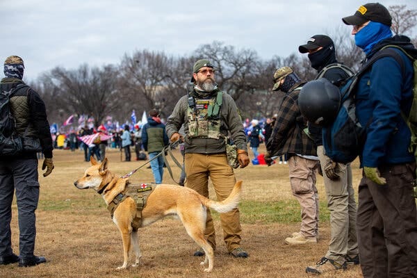 An Oath Keeper with a beard, glasses, a camouflage vest with a two-way radio holds the leash of a dog wearing an Oath Keepers vest while several people with masks, helmets and other tactical gear stand nearby.