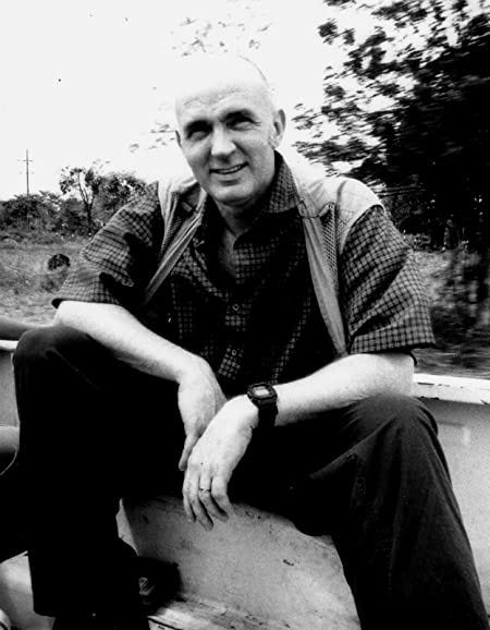 A black-and-white picture of a bald white man in a vest and checkered button-down shirt sitting in nature