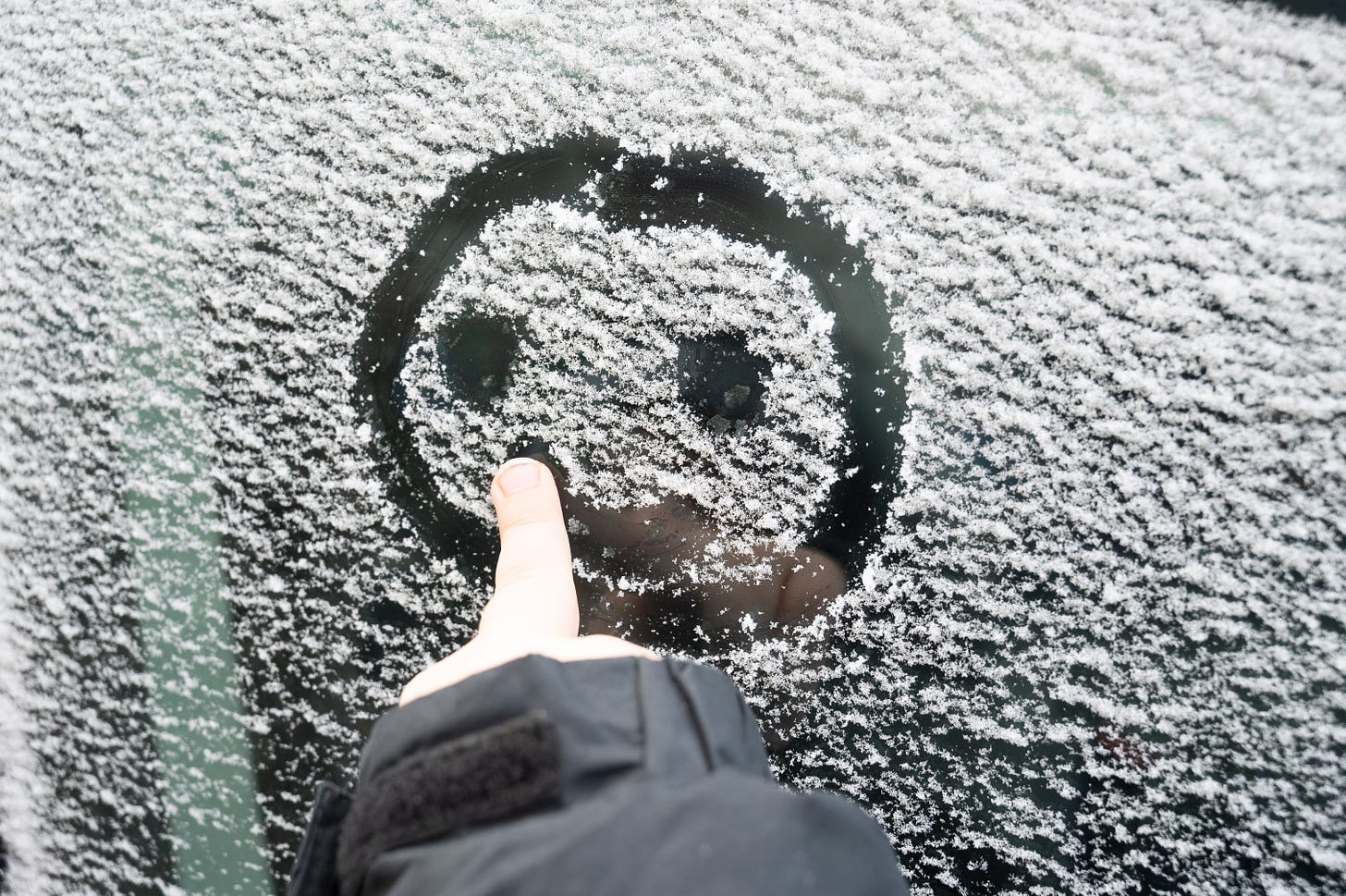 A middle finger poking out of a black coat sleeve drawing a smiley face on a snowy window