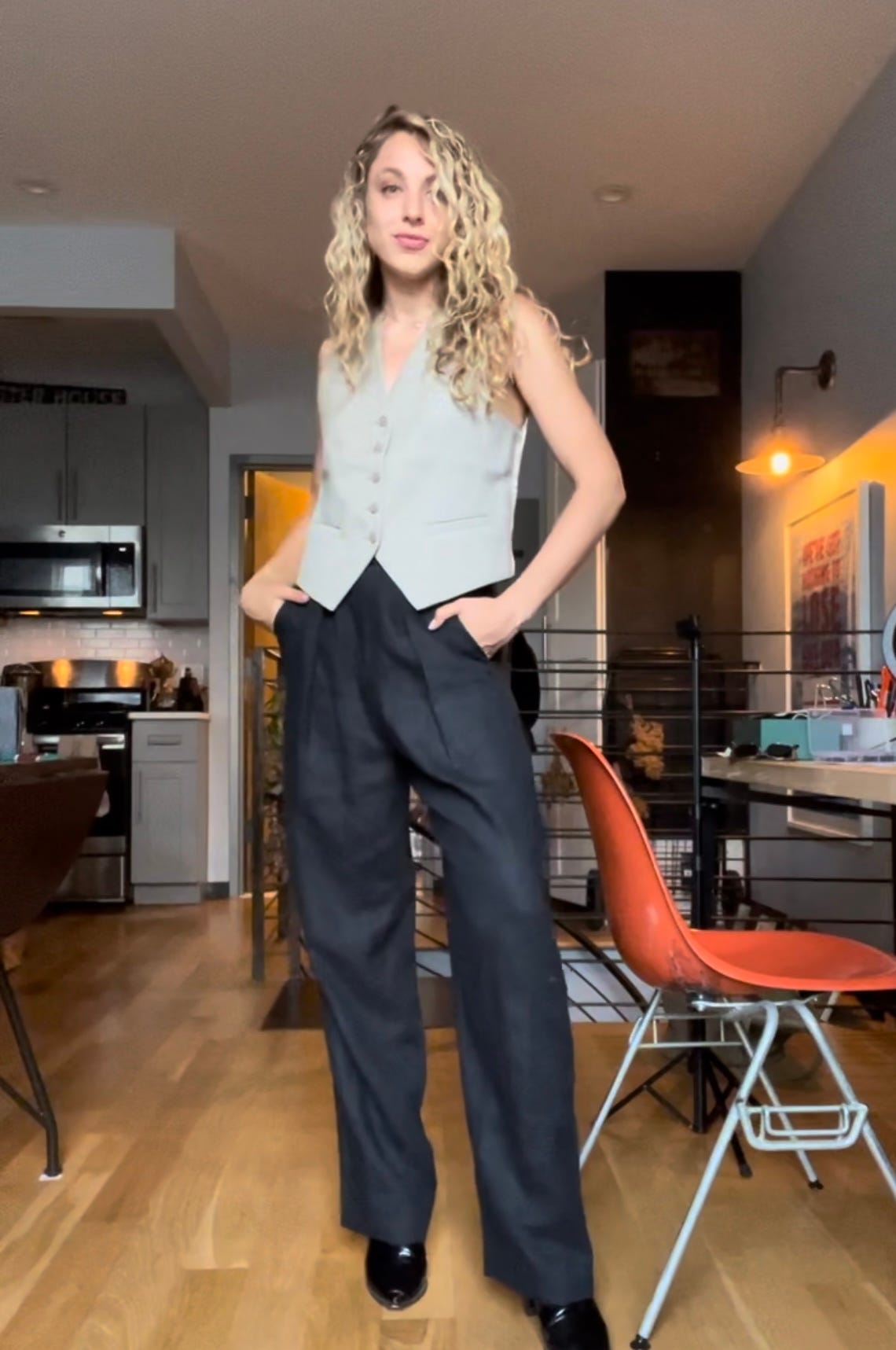 Alyssa is standing in an apartment wearing black pants and a beige vest.