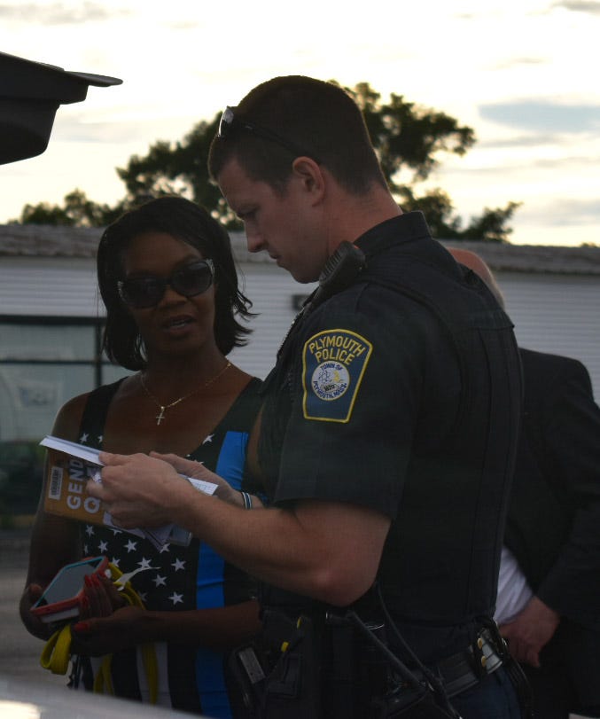 Republican candidate for Massachusetts secretary of the commonwealth Rayla Campbell, wearing a Thin Blue Line dress, speaks with a police officer who is looking through her library copy of Gender Queer to determine if it’s child pornography. (Credit: Rebecca Richards)