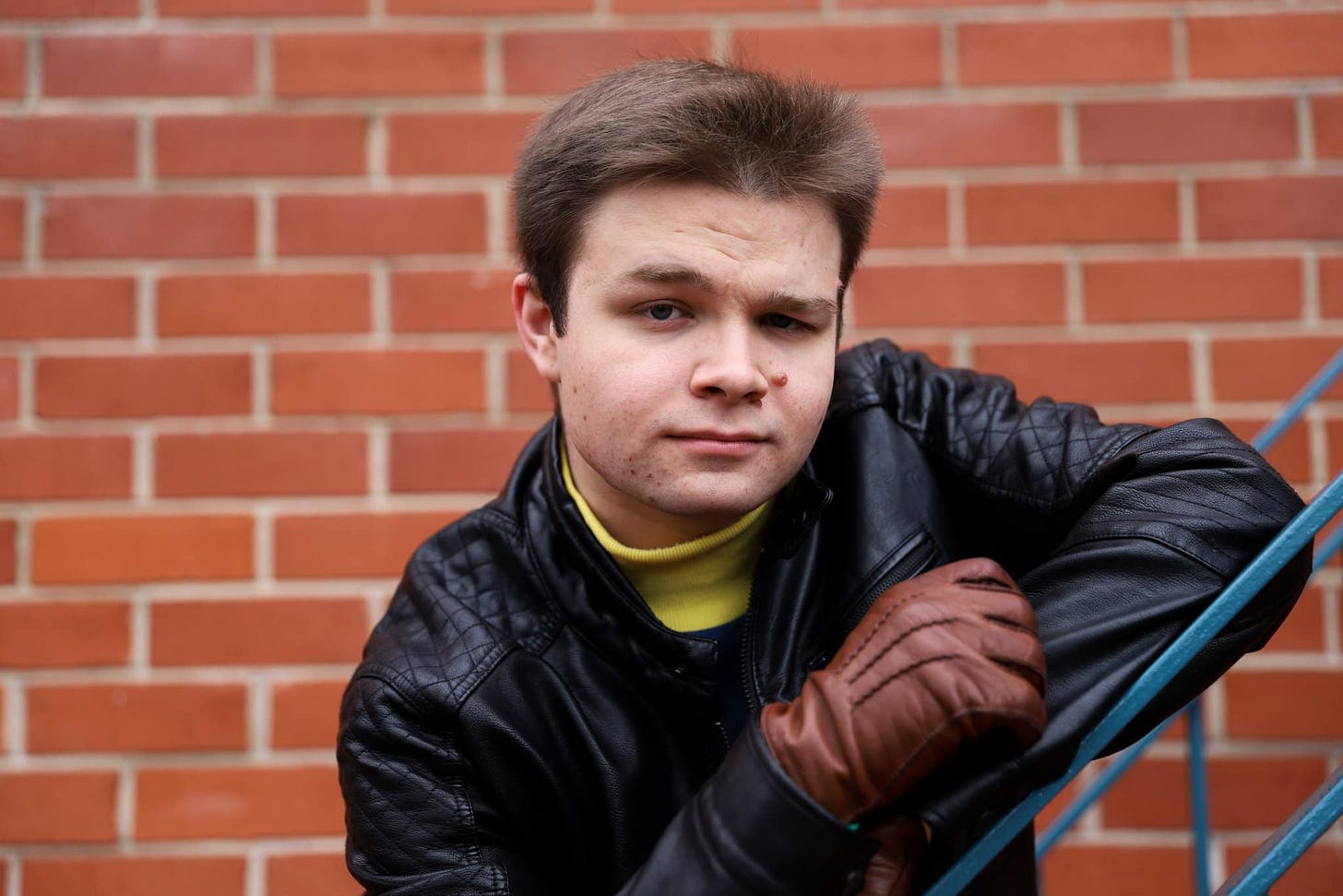 A headshot of John Whinfield. He is leaning on a blue railing against a red brick background. He has short brown hair and is wearing a yellow turtleneck, a black leather jacket and brown leather gloves.