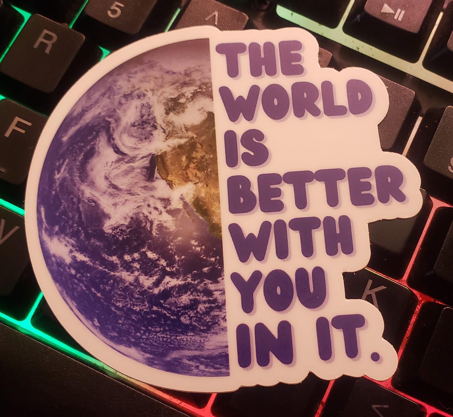 Photo of a sticker that is half a realistic earth with the words The world is better with you in it to the right sitting on a rainbow backlit keyboard. 