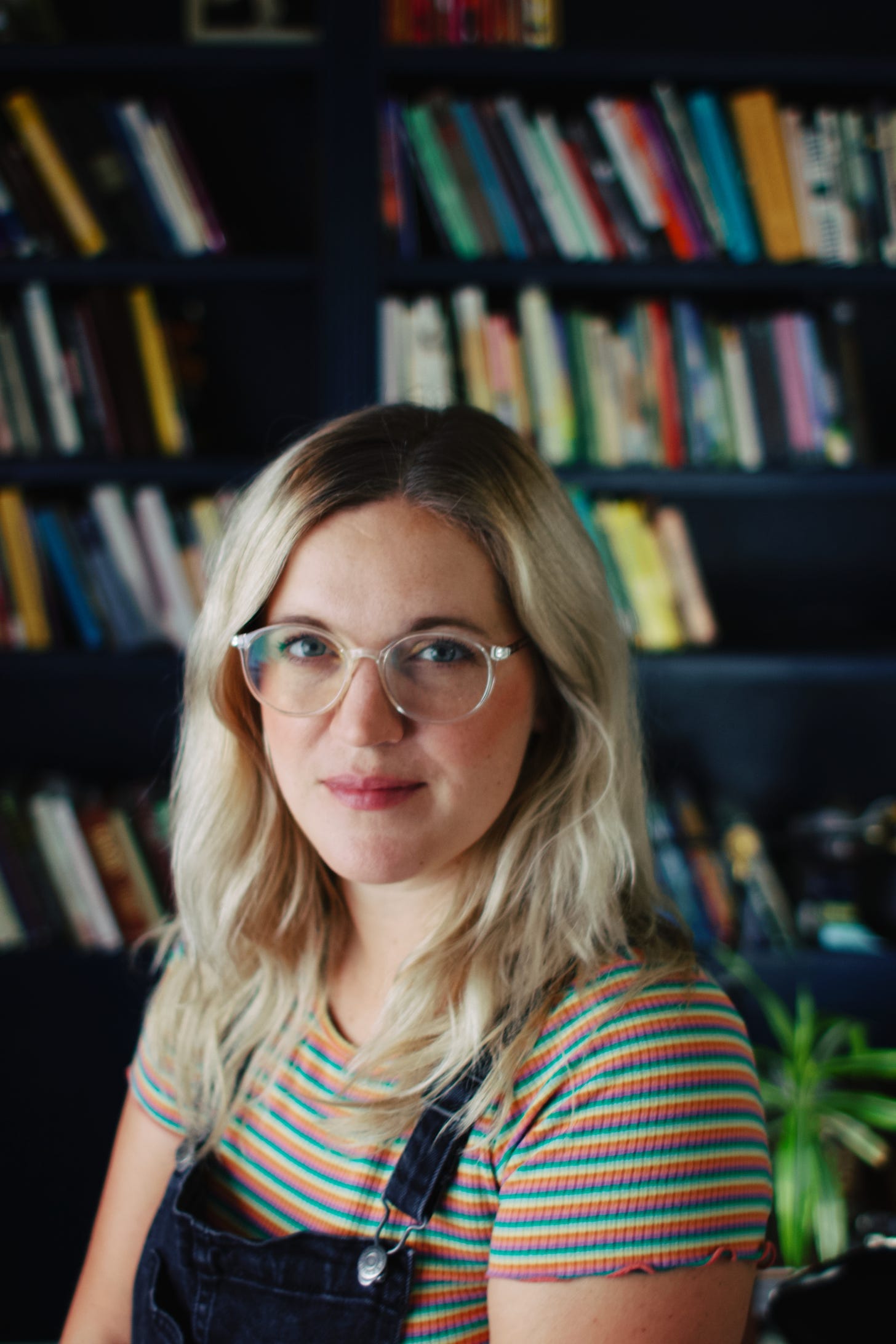 a white woman wearing clear glasses is in front of a dark blue bookcase. she is smiling slightly at the camera, and she has long blonde hair with dark brown roots. she is in her late 30s, and is wearing a rainbow shirt and black overalls