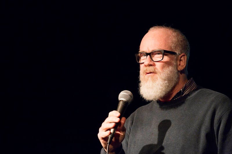 An older bearded man with glasses stands in a spotlight holding a microphone against an otherwise pitch-black backdrop.