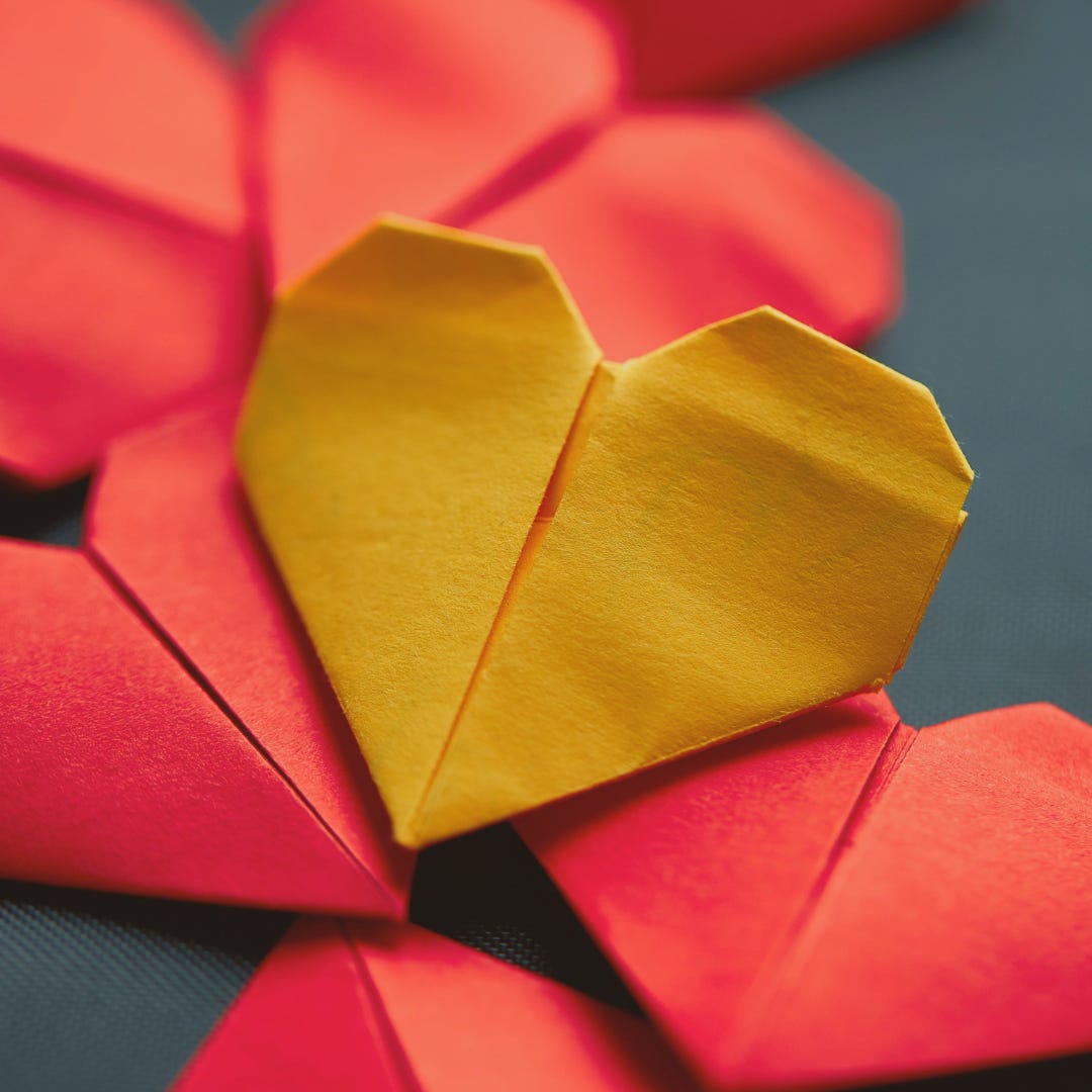 Construction paper has been folder into a bunch a small, sweet paper hearts that have been scattered across a wooden table. 