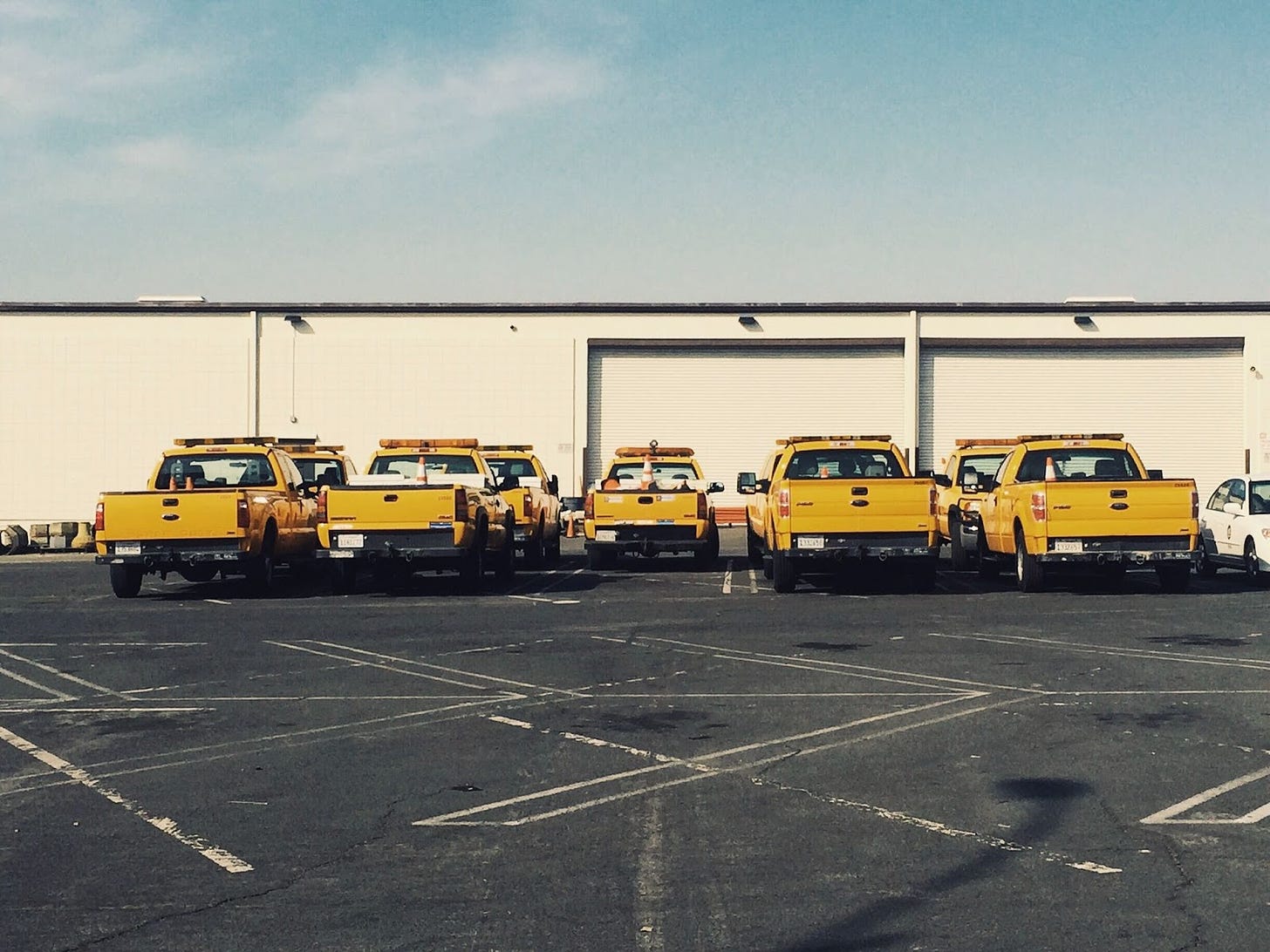 Photo of nine yellow pick up trucks parked next to one another with asphalt and parking spot lines in the foreground and the Bureau of Street Lighting in the background.