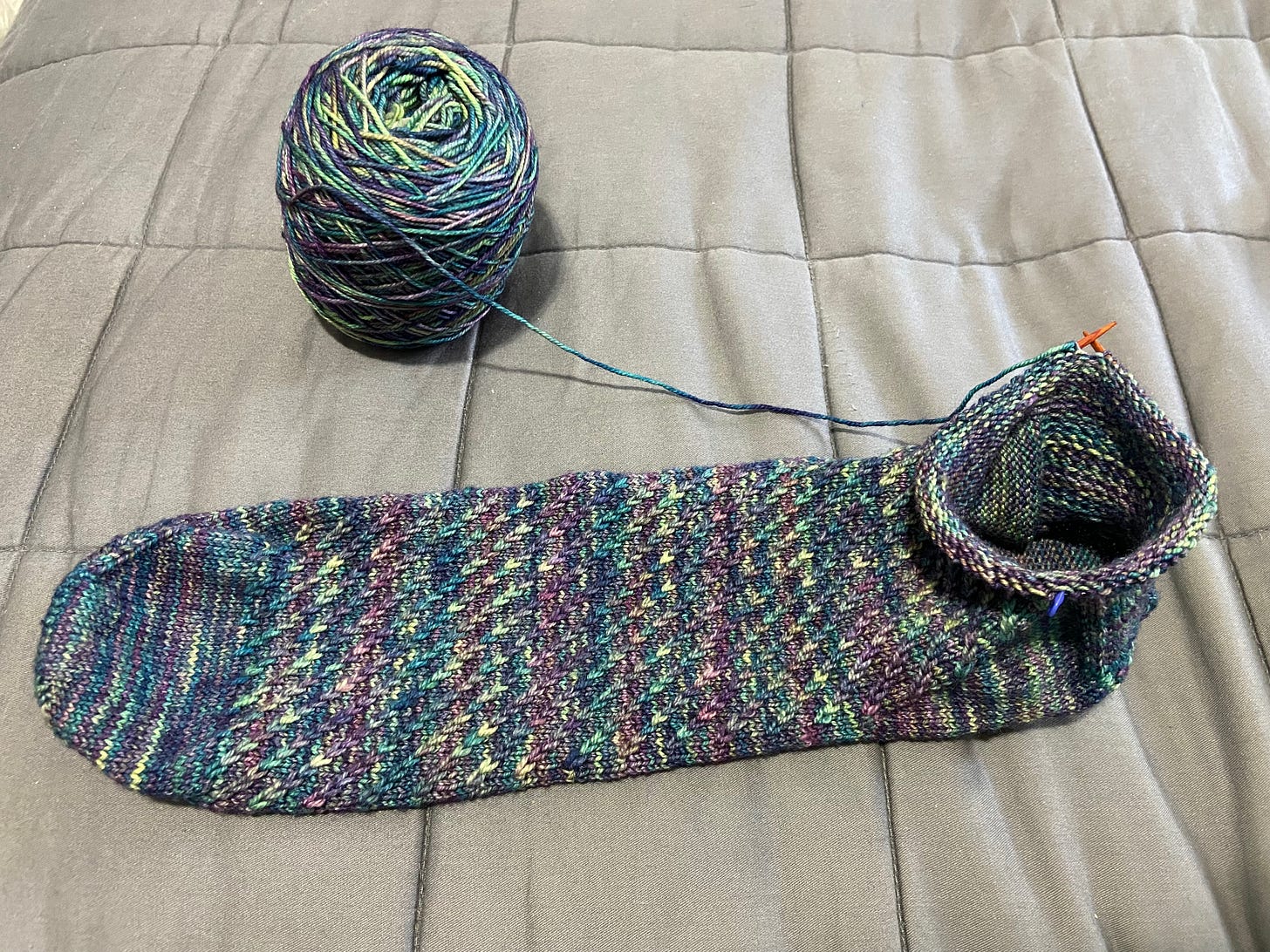 A green, blue, and purple sock (still in-progress) knitted toe-up is displayed on a bed; I just started on the leg. Tiny circular needles and a partial skein are attached.