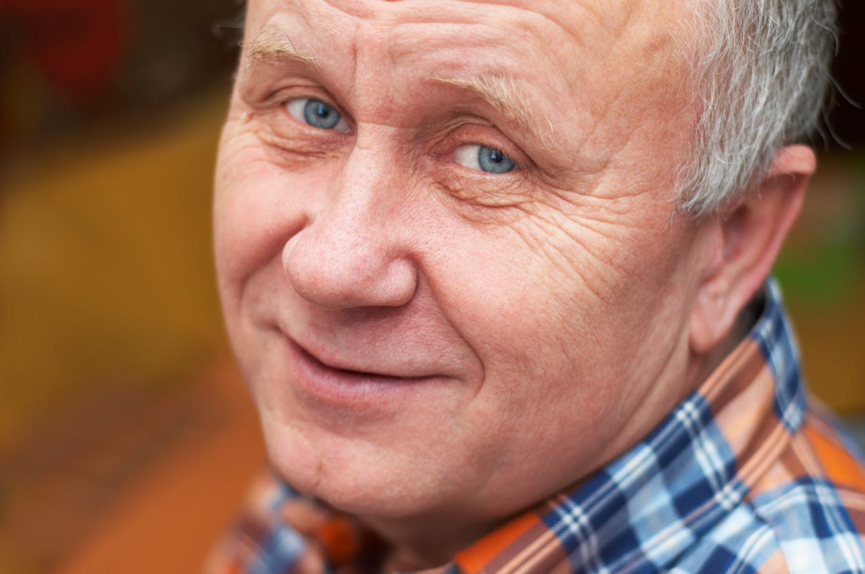 Tight facial photo of white, aging man with graying hair, blue eyes, no glasses and troubled smile.