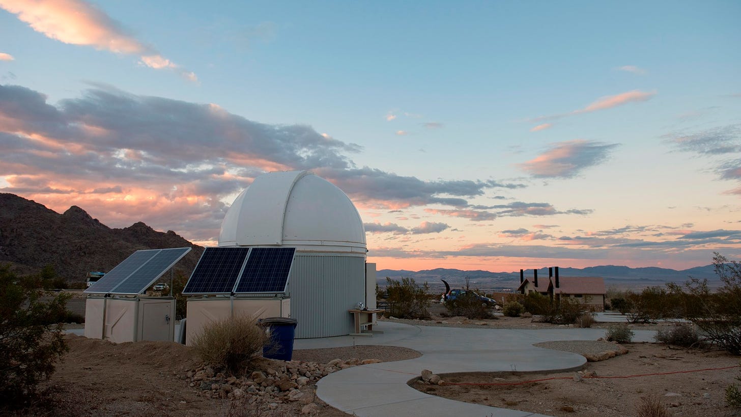 A 15 foot observatory with a paved path in front of clouds lit by sunset over mountains.