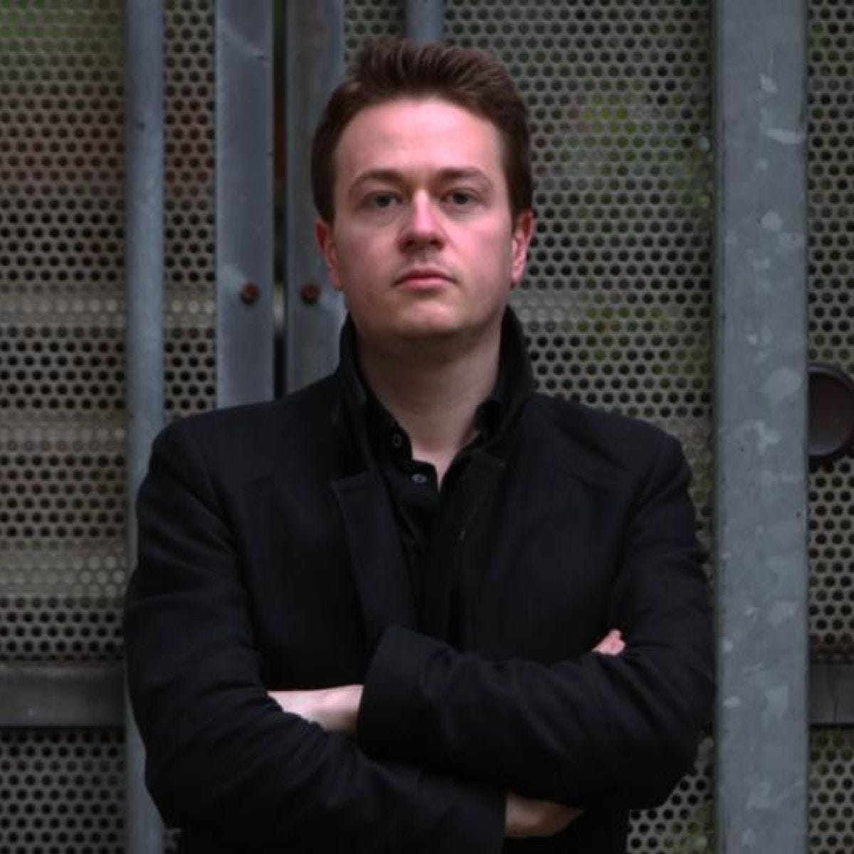 Johann Hari standing in front of a metal fence with his arms crossed