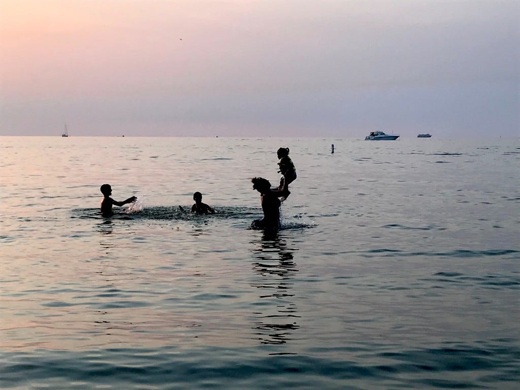 a woman stands waist deep in a calm Lake Erie as the sun sets, silhouetting her against a pale purple sky and the silver rippling water surface. she playfully tosses a young child into the air as two other children splash each other behind her.