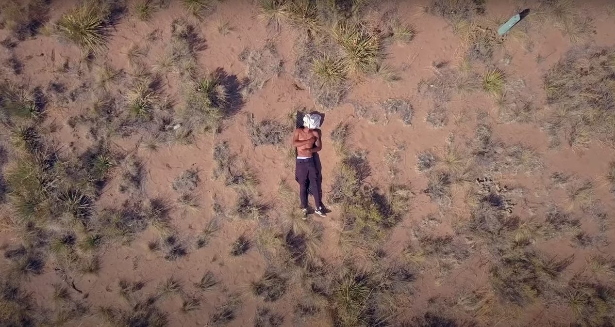 A camera looks to the ground from above at a shirtless man, laying face up, resting in the Colorado nature. From the film 'Crestone'.