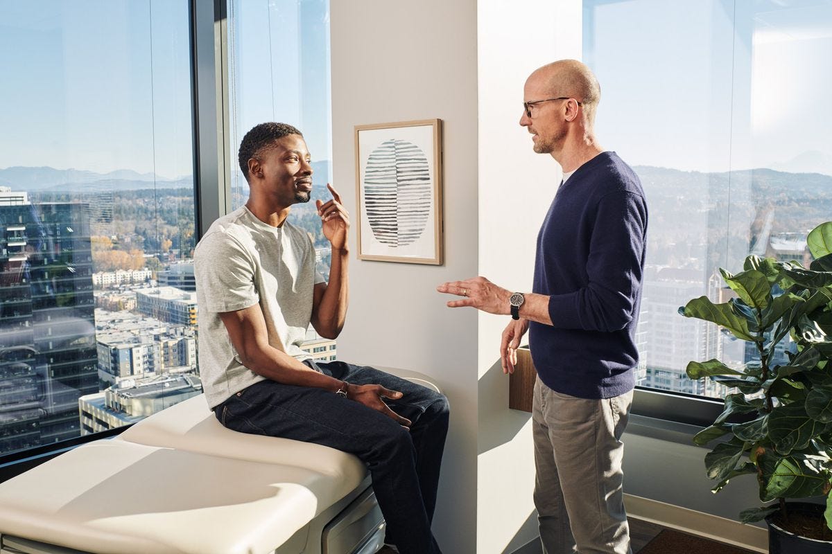 In a promotional image from One Medical, a doctor that looks just like REM singer Michael Stipe talks to a young Black man sitting on a doctor’s office bed in a sun-flooded high-floor glass walled corner office. In a promotional image from One Medical, a doctor that looks just like REM singer Michael Stipe talks to a young Black man sitting on a doctor’s office bed in a sun-flooded high-floor glass walled corner office.