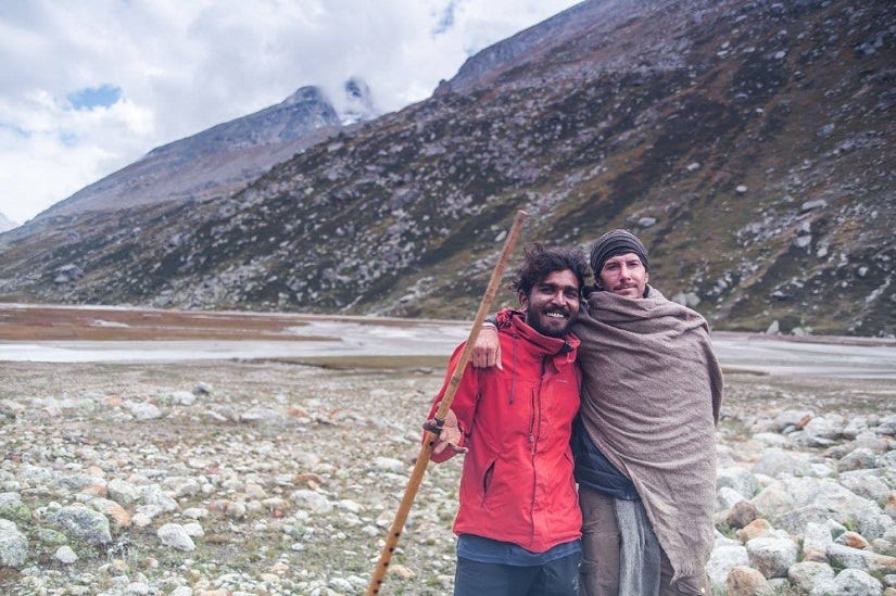 A man in a red jacket stands next to Justin, who is wrapped in a blanket, with mountains behind them.
