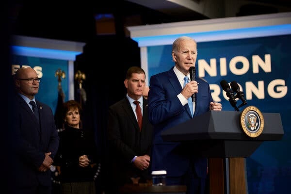President Biden holds a microphone up while standing in front of a blue sign that says “union strong.”