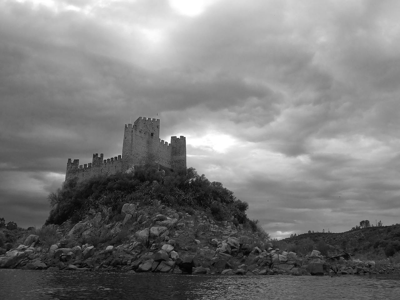 A small castle sits on a rocky hill surrounded by water, with heavy gray clouds above.