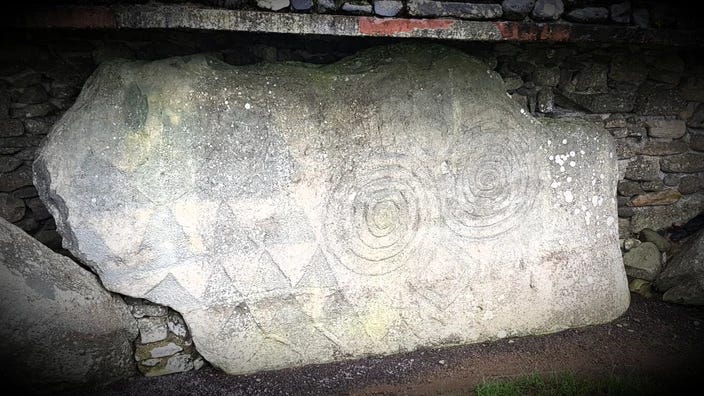 A large grey roughly rectangular stone with several rows of picked triangles to the left and two striking concentric circular motifs centre and right surrounded by smaller diamond shaped carvings