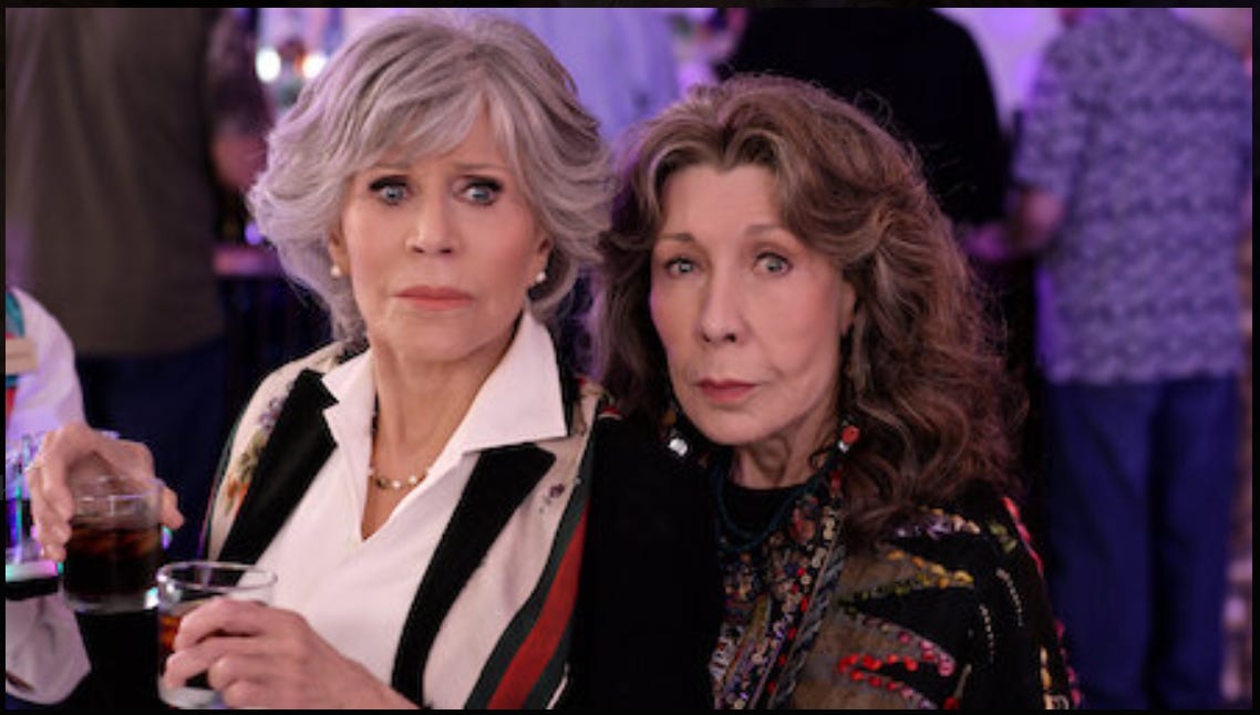 Grace e Frankie, interpretate da Jane Fonda e Lily Tomlin, in una scena della serie: le due donne guardano verso di noi con uno sguardo allarmato. Grace regge tra le mani due bicchierini di liquore.