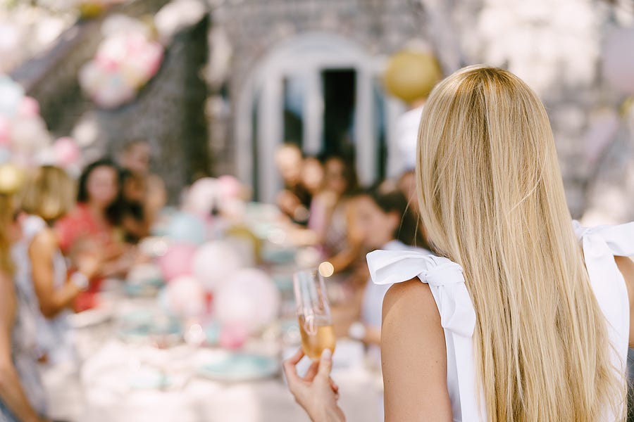 A blonde woman in a white sleeveless dress with a glass of champagne in her hand stands in the foreground with her back to the camera, looking at a table at a wedding banquet which is out of focus in the background