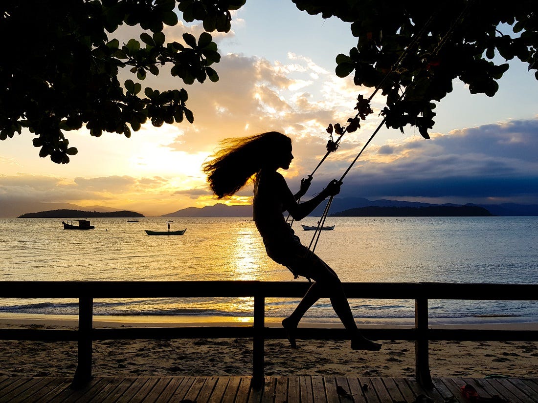 Foto de uma menina de uns 12 anos num balanço ao pôr do sol com o mar alaranjado atrás dela.
