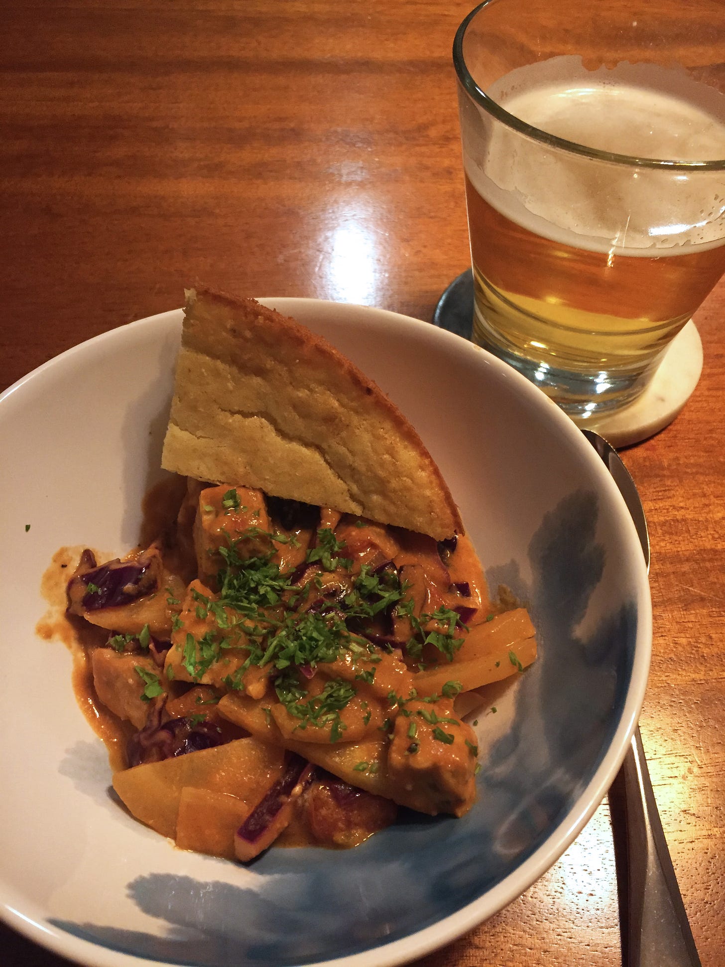 In a white bowl, an orangeish stew with visible pieces of carrot and cabbage is topped with parsley. A wedge of cornbread rests at the side of the bowl. Beside it on a coaster is a glass of lager.