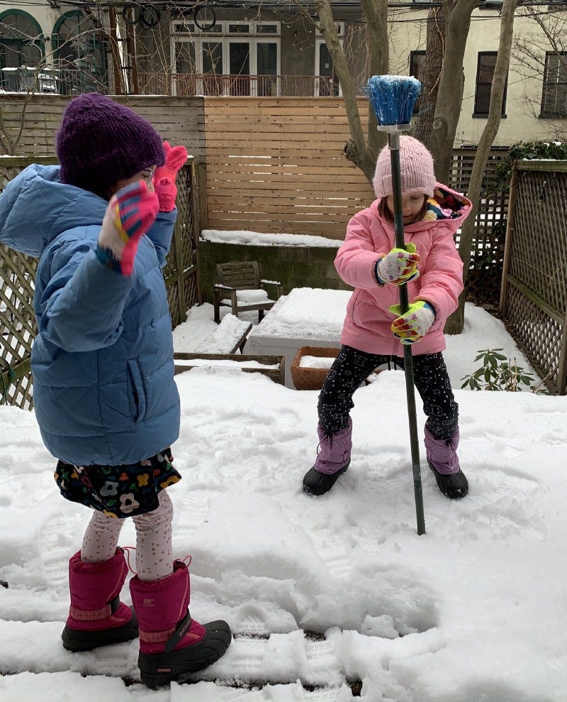 two kids play out in the snow. one jams a broom handle into the snow while the other watches.