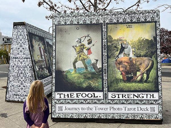 A photo of two large freestanding walls on a street corner that have giant tarot cards wheat-pasted onto them as an installation for something called Journey to the Tower Photo Tarot Deck, by artists Two by Sea. These are a photographic interpretation of the classic Rider Smith Waite deck. In this photo, the back of a kids head can be seen as they look at The Fool and Strength. A tent with a blue tarp thrown over it is visible in the background. 