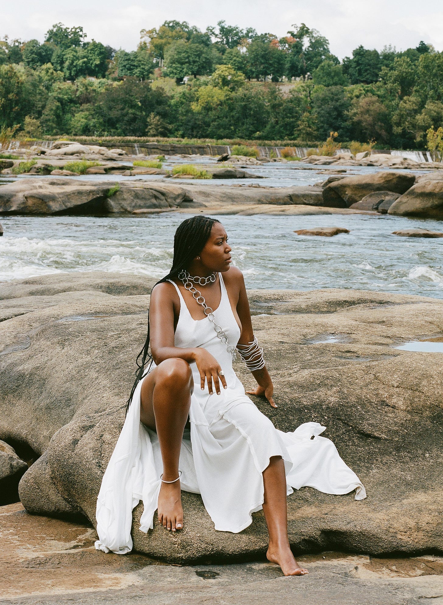A black woman (me) with braids wearing a white dress looking away from the camera while sitting on top of river rocks with a tree line in the background.