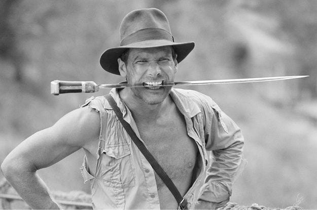 Indiana Jones with a giant sword in his teeth, one sleeve ripped and his shirt open.