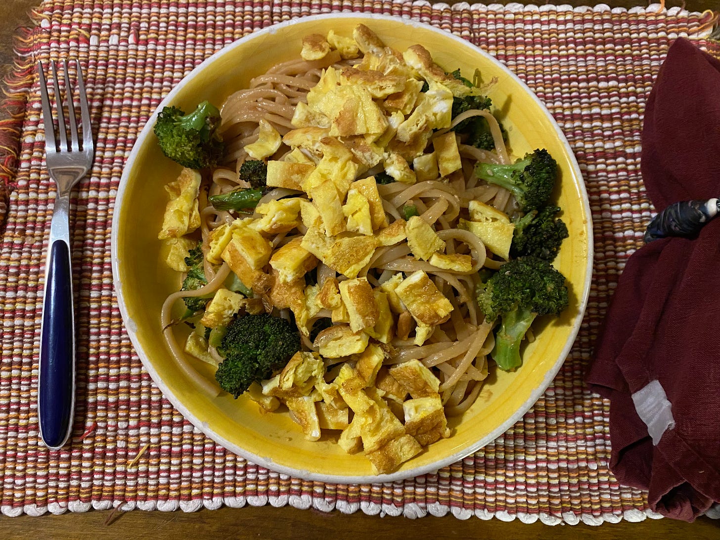 A wide yellow bowl sits on a woven placemat, a fork on one side and a red folded napkin on the other. The bowl is full of noodles, topped with roasted broccoli and small squares of cut-up omelette.