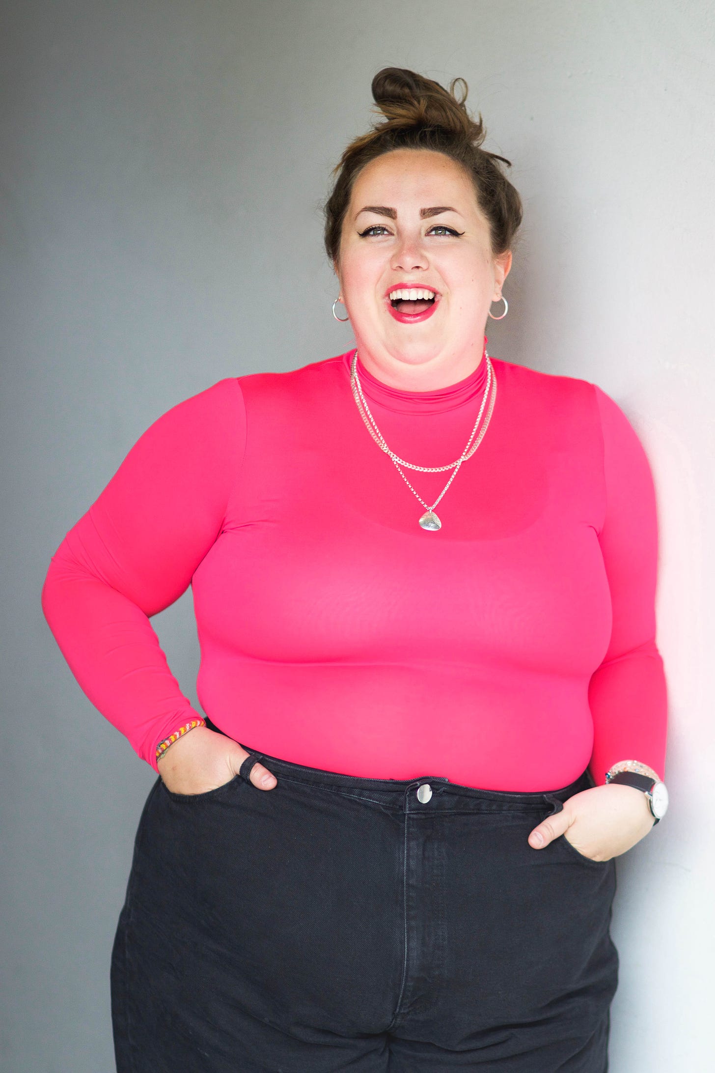 Headshot of Katie Greenall, wearing a pink turtleneck jumper, standing against a blank wall