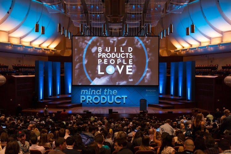 Foto mostra um auditório cheio de pessoas sentadas, um palco vazio e um telão, em que está escrito: "Build Products People Love. Mind The Product" ("Construindo produtos que as pessoas amam. Mind The Product").