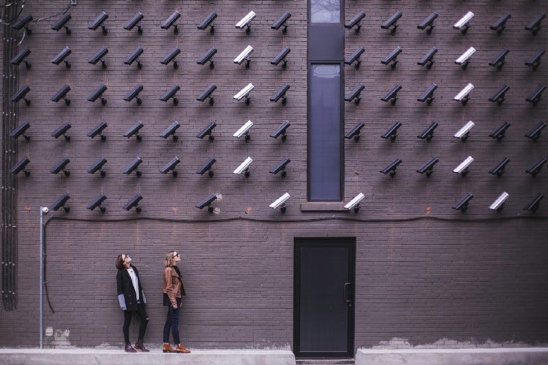 Two women looking up at a brick wall of fifty CCTV cameras.