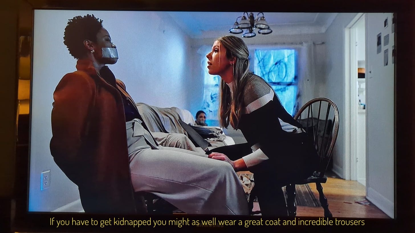 Victoria, wearing a beautiful long red coat and wide-leg paperbag trousers, tied to a chair while Claire holds a knife toward her. Captioned "if you're going to be kidnapped you might as well wear a great coat and incredible trousers"