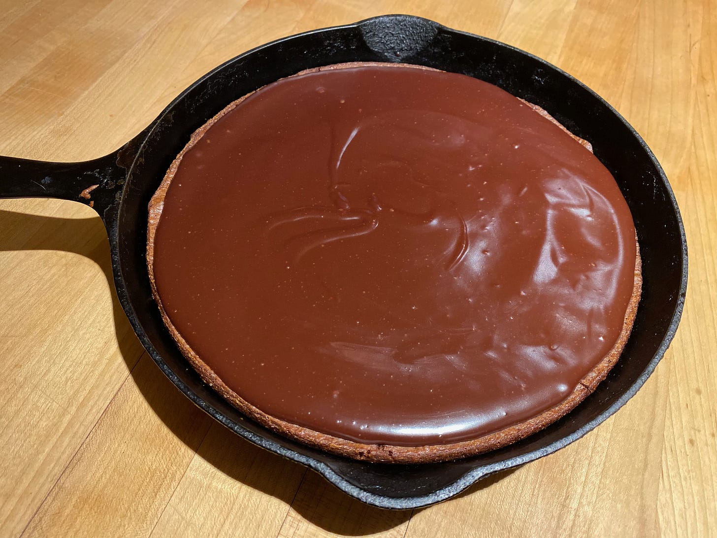 A cast iron pan holds a large round brownie, topped with a shiny chocolate ganache.