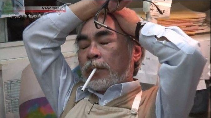 Cena do documentário, com Miyazaki, homem japonês de barba branca, camisa social e e avental, tirando os óculos e passando a mão na cabeça de olhos fechados. Na boca, um cigarro apagado.