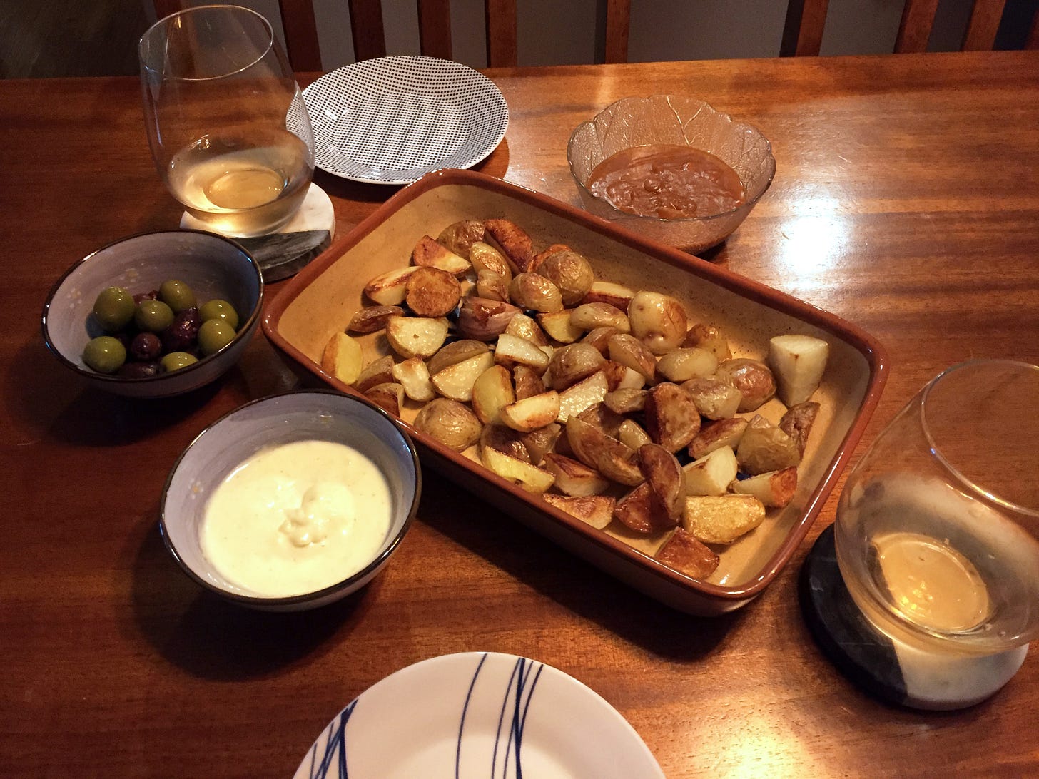 A shallow rectangular dish full of browned wedges of potato, surrounded by smaller dishes with olives, garlic mayo, and salsa brava. Two glasses of rosé wine sit on coasters next to two small empty plates.