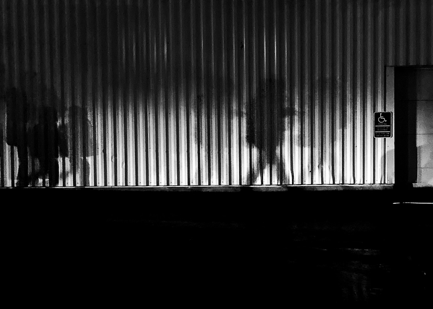 A corrugated metal wall outside of the stadium shows the shadows of the passing people illuminated by car lights.