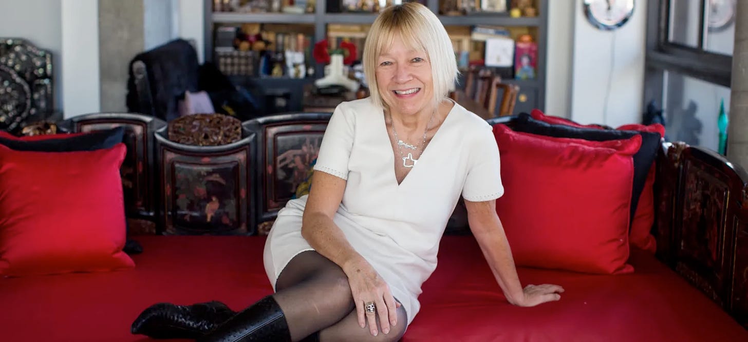 Photo of Cindy Gallop in a white dress and black boots, seated on a red couch, smiing