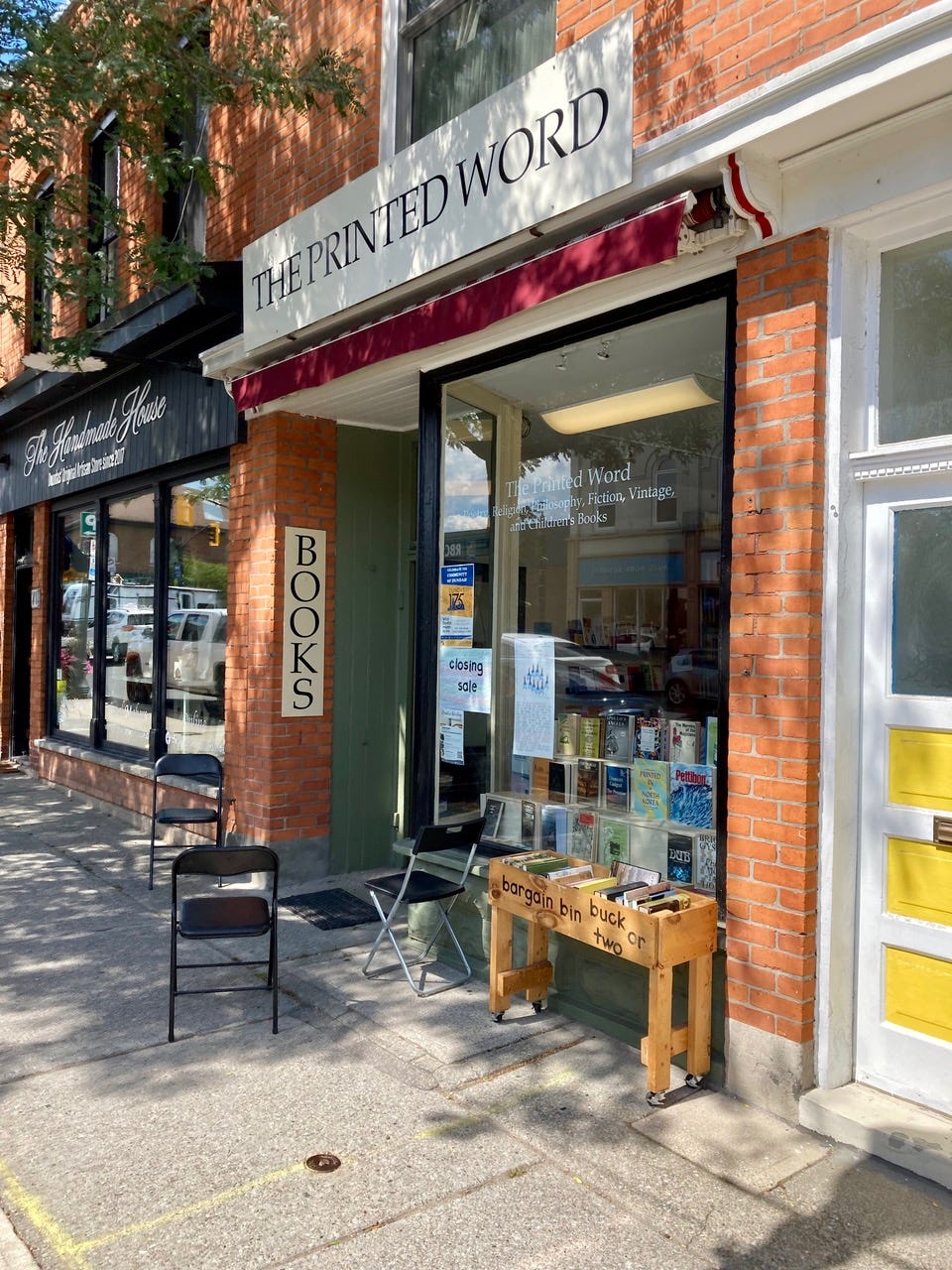 Exterior of a red brick building with a glass storefront filled with books on display. A sign above reads The Printed Word. Three folding chairs are set up beside a wooden cart with black lettering: bargain bin, buck or two