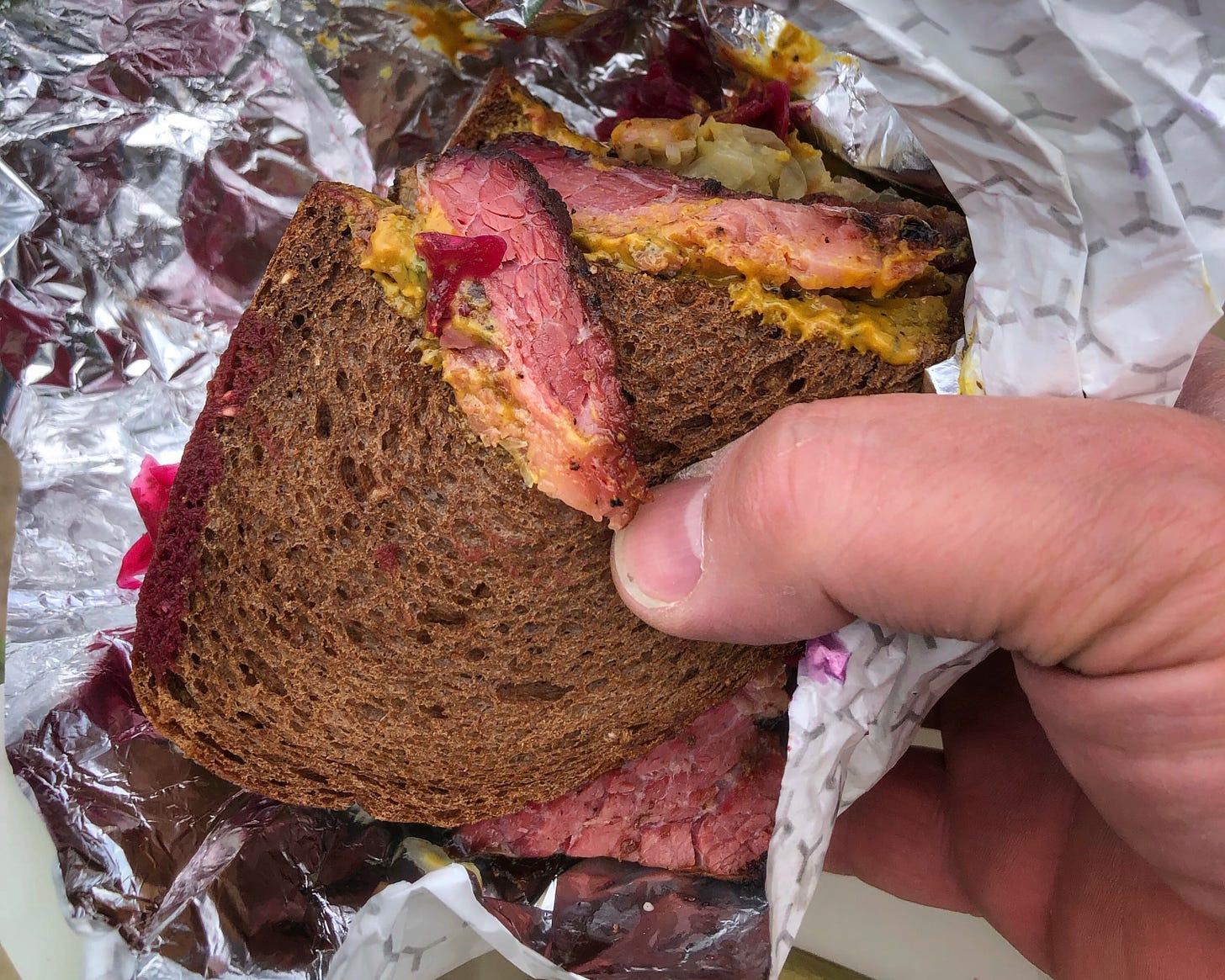 A hand holding a thick-cut pastrami sandwich on pumpernickel. There are visible patches of yellow mustard and red cabbage sauerkraut.