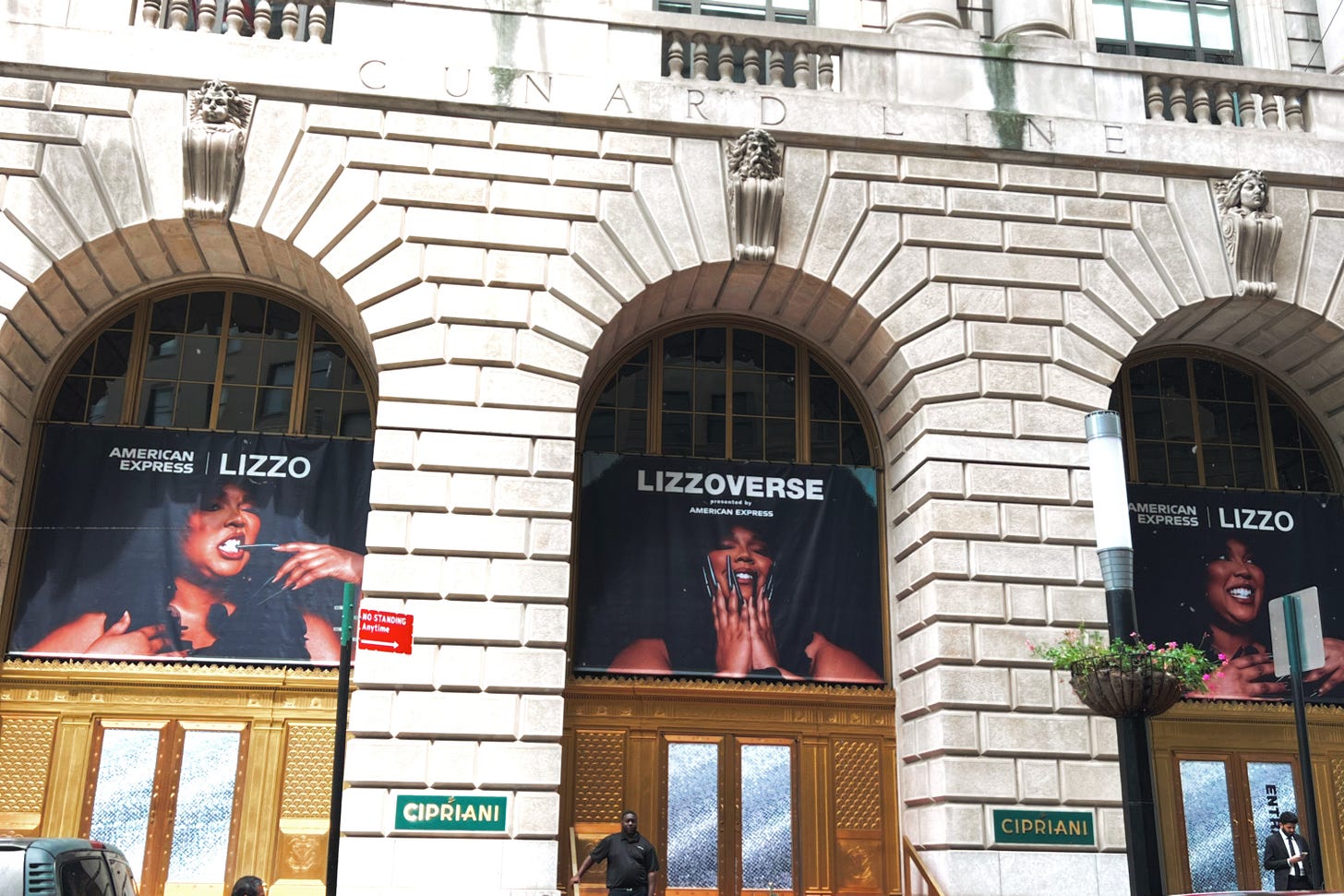 A photo of Cipriani 25, a white stone building with three archways and three posters of Lizzo hung in each one.  Below each poster is a gold art-deco style doorway with speckled, frosted glass panes in the door.